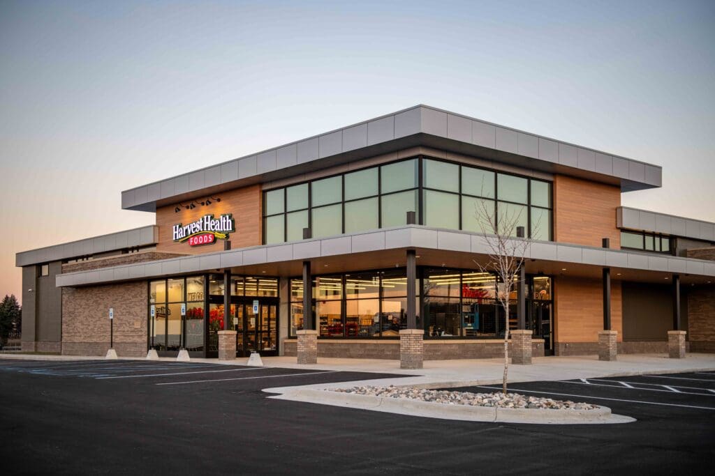 A modern, single-story grocery store called Harvest Health Foods, with large windows and a mostly empty parking lot, photographed at dusk.