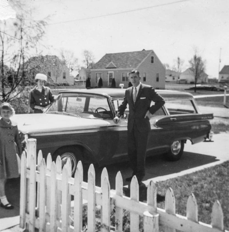 Black and white photo of a man in a suit, a woman in a hat, and a young girl standing by a classic station wagon near a white picket fence in a suburban neighborhood, with houses in the background.