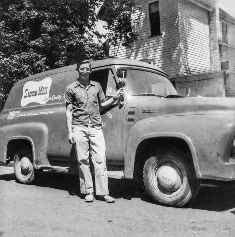 A man stands smiling beside a vintage delivery van labeled Stone Mill, holding a trophy. The scene is outside, with a large house and trees visible in the background.