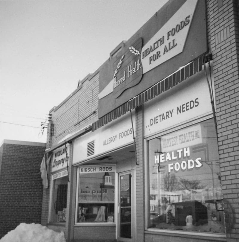 Black-and-white photo of a storefront named Harvest Health with signs reading HEALTH FOODS FOR ALL, ALLERGY FOODS, DIETARY NEEDS, and HEALTH FOODS. A pile of snow is visible in the foreground.