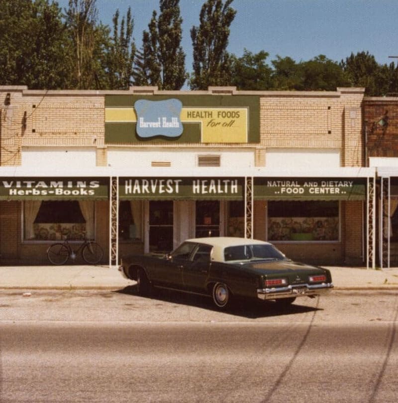 A vintage car is parked in front of a brick storefront called Harvest Health, which sells vitamins, herbs, books, and natural foods. A bicycle is parked to the side, and trees are visible in the background.