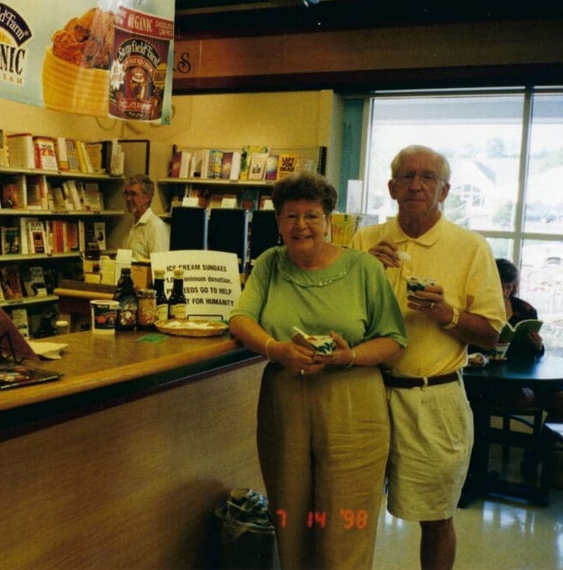 An older couple stands and smiles in an ice cream shop, holding cups of ice cream. Behind them is a counter with books and a man working. Sunlight streams through large windows. The date 7 14 98 is on the photo.