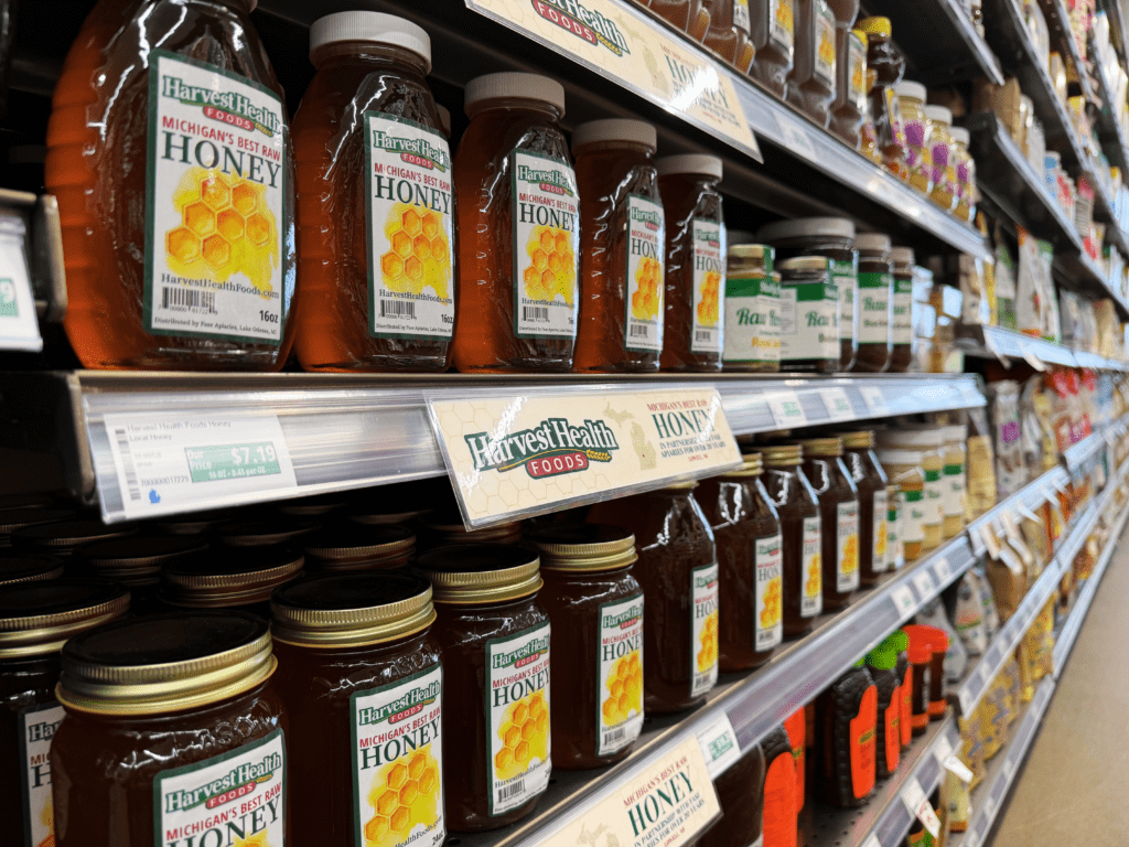 Shelves in a grocery store stocked with multiple jars and bottles of Harvest Health Foods raw honey, with price tags and product signage displayed along the aisle.