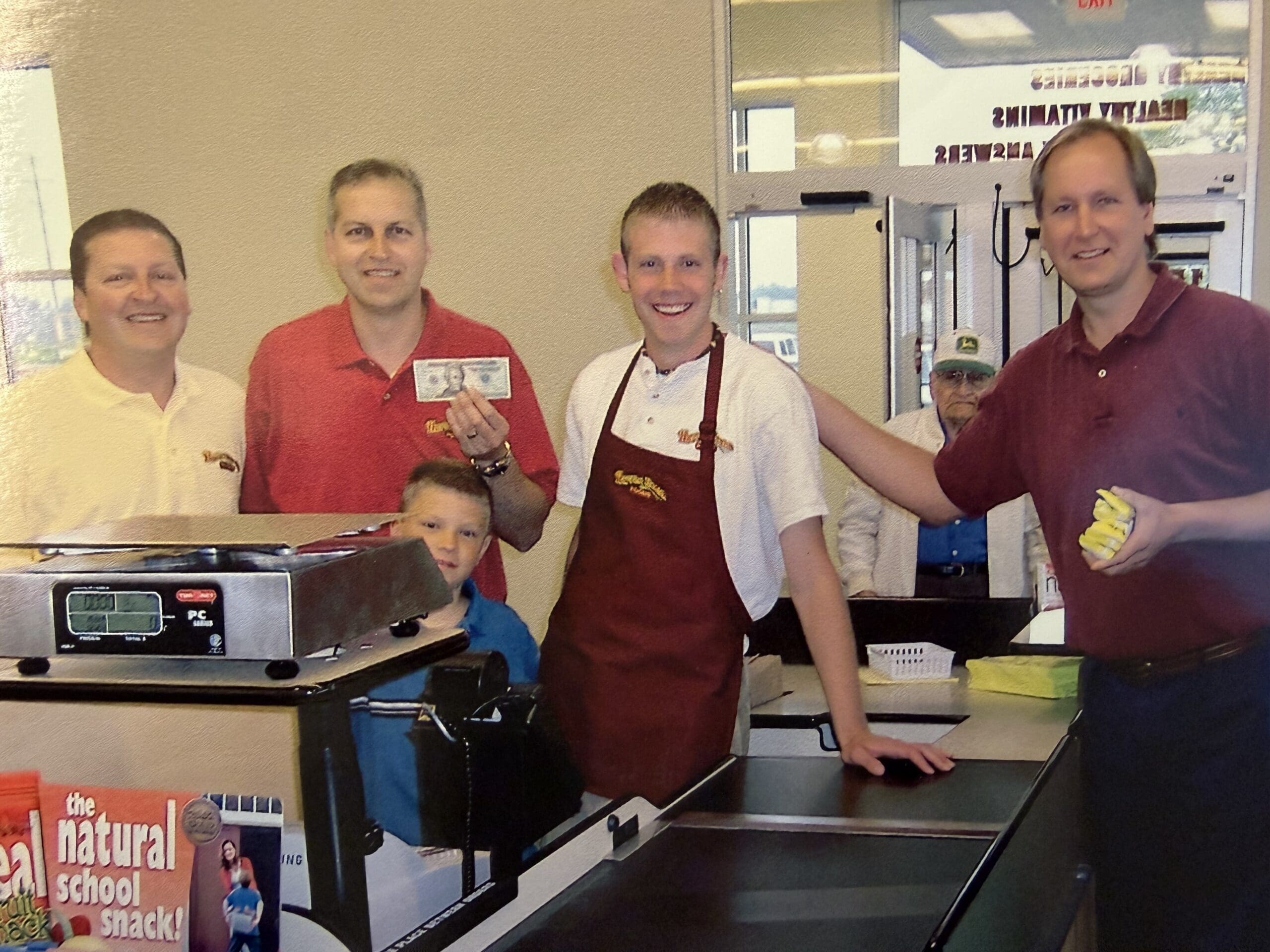 A group of five men and a child smile for a photo at a grocery store checkout counter; one man holds up cash while another wears an apron behind the counter. A person can be seen entering through the glass door in the background.